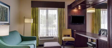 Habitación de hotel con sofá verde, un escritorio, una silla, un reposapiés, un televisor de pantalla plana y un ventanal con cortinas.