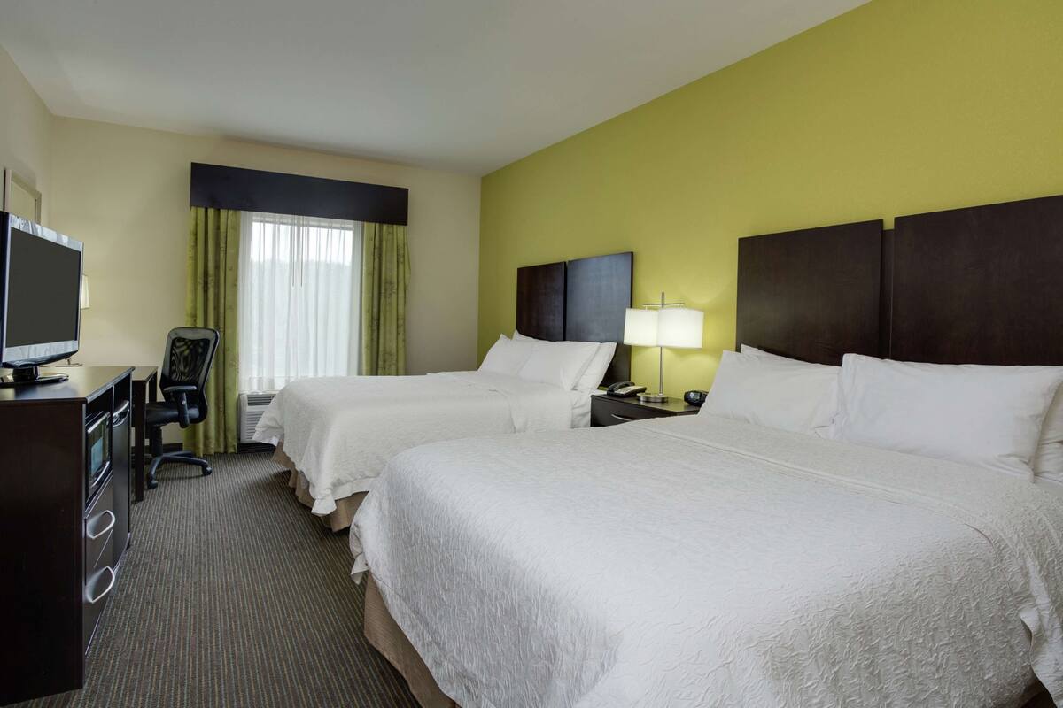 A hotel room with two beds, a desk, a chair, a TV, and a window with curtains.