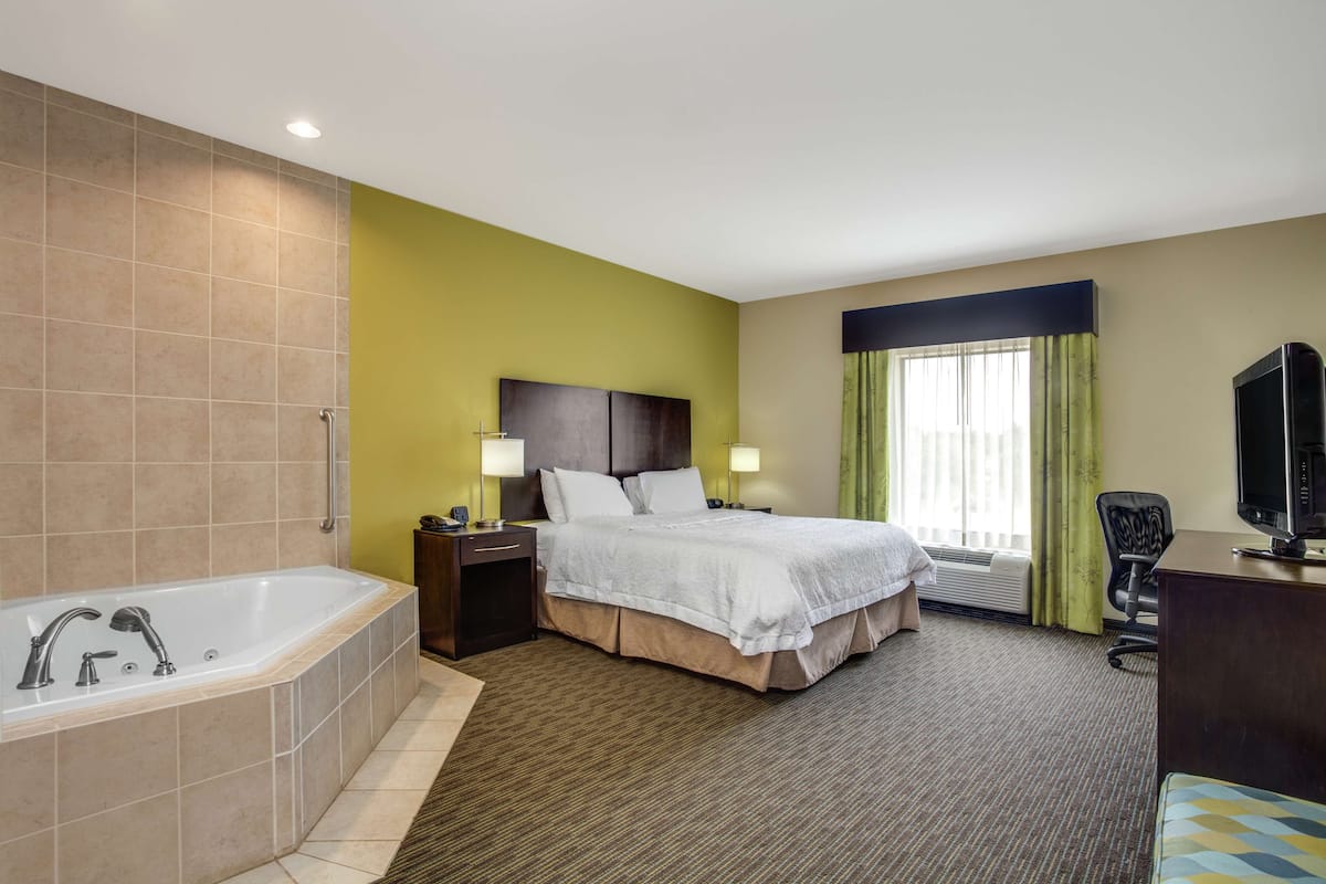 A hotel room with a large bathtub, a bed with white linens, a desk with a television, and a window with curtains.