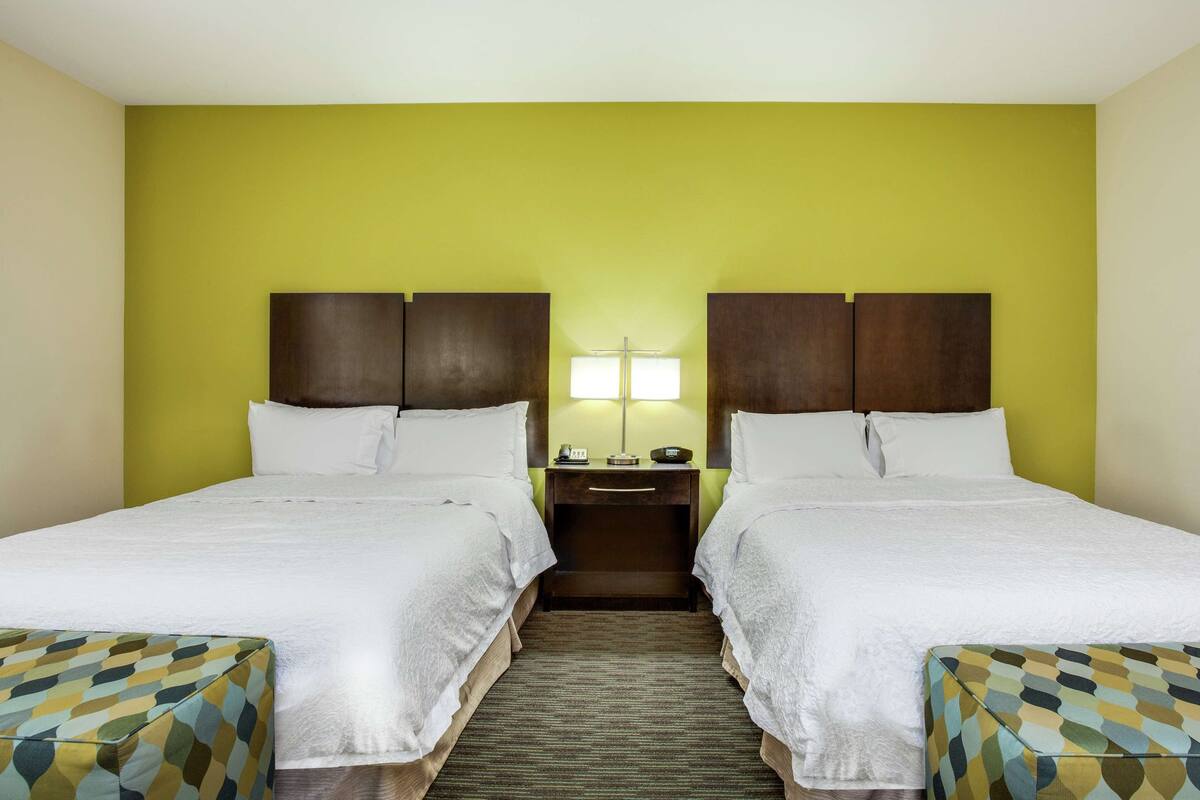A hotel room with two beds, a nightstand with a lamp, and a patterned ottoman.