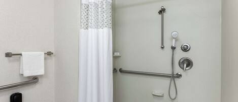 Zona de ducha con puerta de vidrio, cortina de ducha blanca, ducha de mano y barra de apoyo.