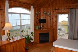 A wooden cabin interior with a fireplace, a television mounted above it, a dining area with a table and chairs, and a large window with curtains.