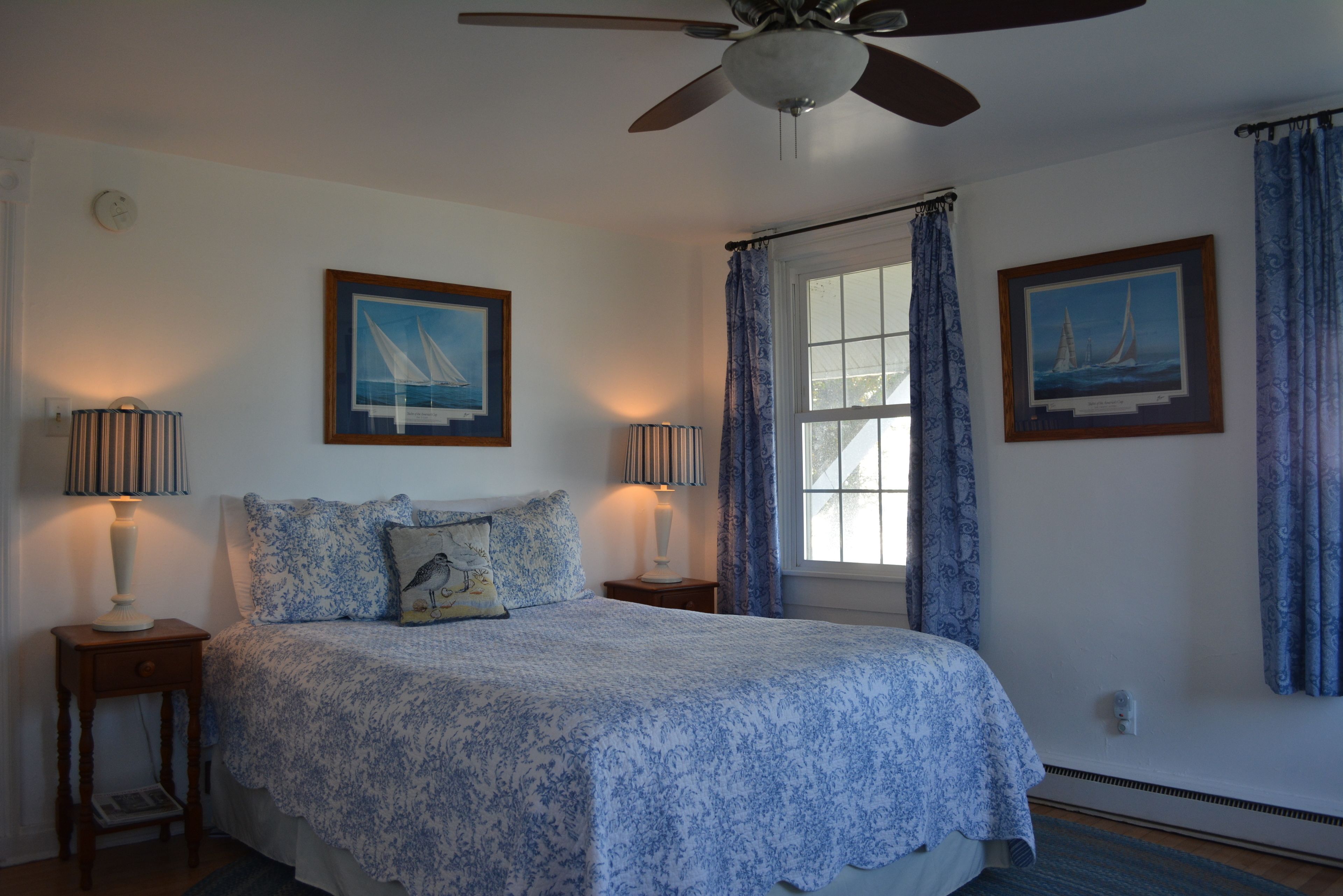 A bedroom with a bed, two bedside tables, a ceiling fan, and two framed pictures on the walls.