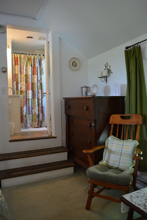 Une pièce comprenant une chaise en bois, une petite table, un meuble et une fenêtre ornée d’un rideau à motifs floraux.