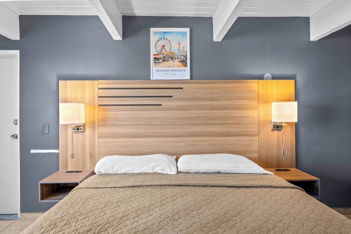 A hotel room with a wooden headboard, two bedside lamps, a bed with a beige quilt, and a poster of a seaside scene.