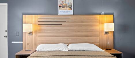 Quarto de hotel com cabeceira de madeira, duas luminárias de cabeceira, cama com colcha bege e um quadro com cena de praia.
