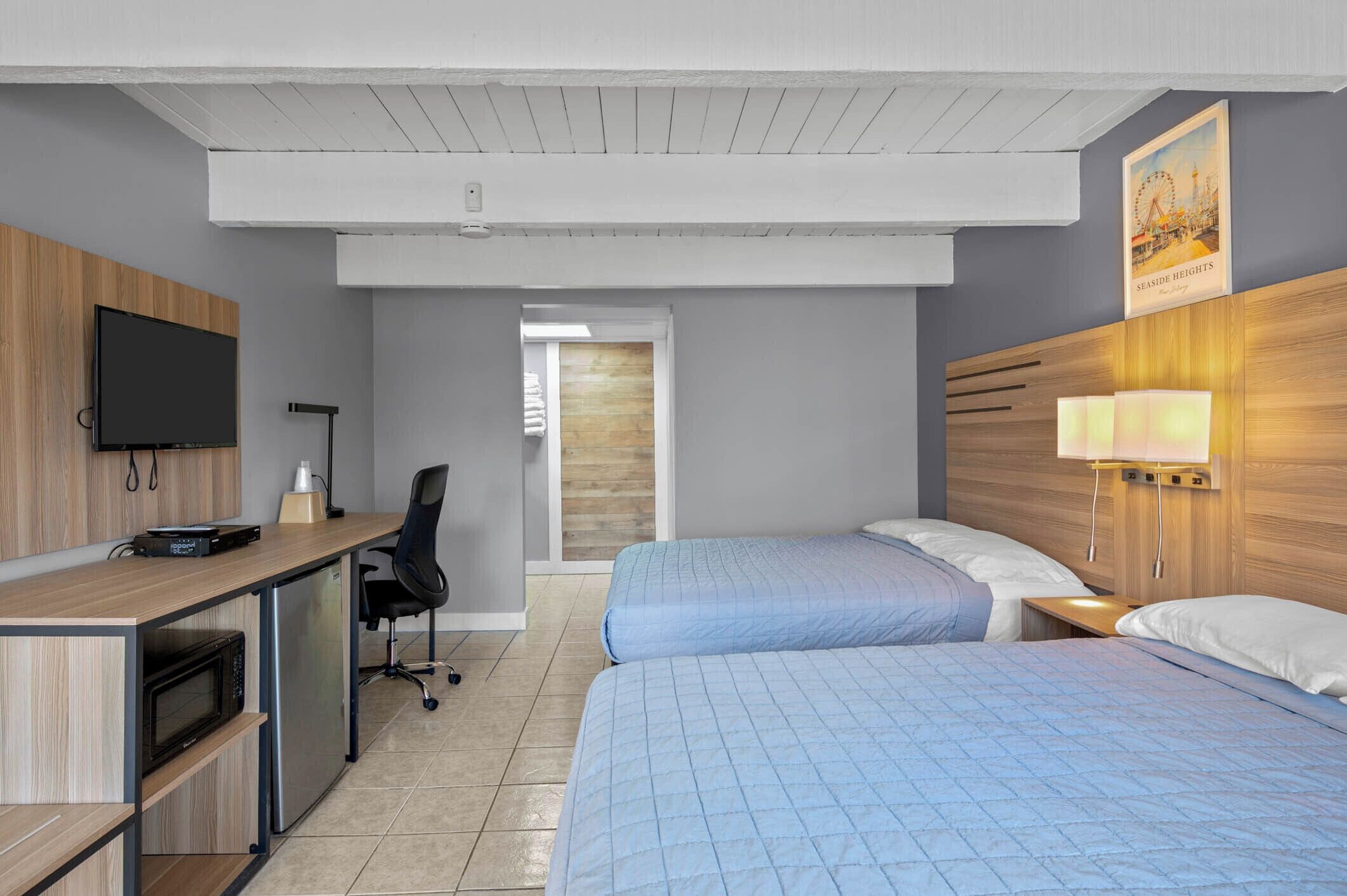 A hotel room with a wooden desk, a flat-screen TV, a bed with blue bedding, a nightstand with a lamp, and a poster on the wall.