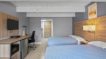 A hotel room with a wooden desk, a flat-screen TV, a bed with blue bedding, a nightstand with a lamp, and a poster on the wall.