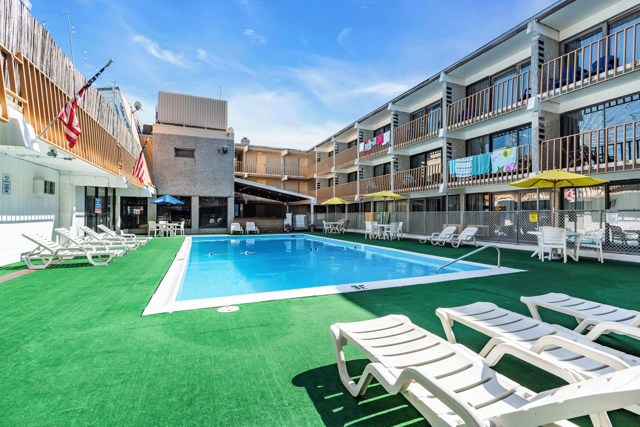 A swimming pool with sun loungers and umbrellas, surrounded by a building with balconies and a covered area.