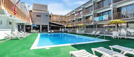 Una piscina con tumbonas y sombrillas, rodeada por un edificio con balcones y una zona cubierta.