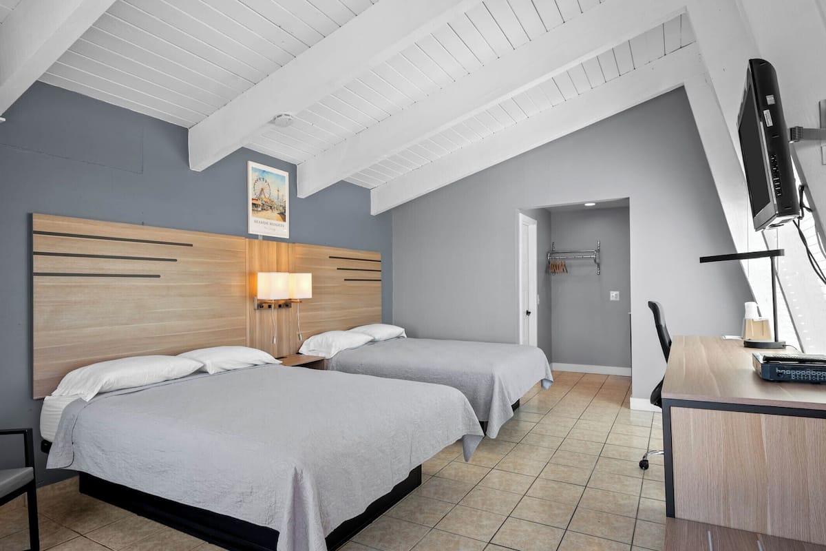 A hotel room with two beds, a desk with a computer, a TV, and a poster on the wall.