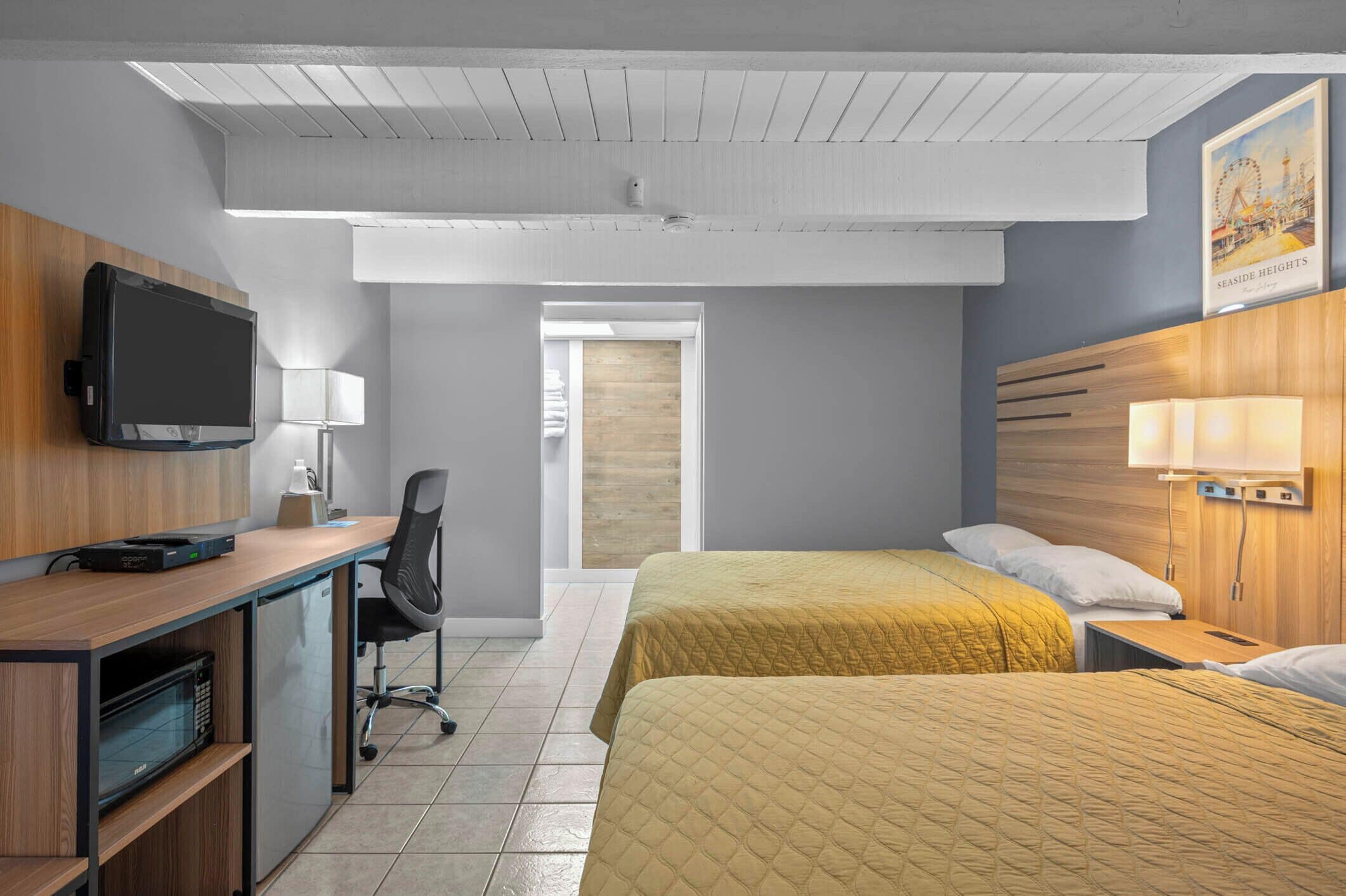 A hotel room with a wooden desk, a television, a bed with yellow bedding, a nightstand with a lamp, and a poster on the wall.