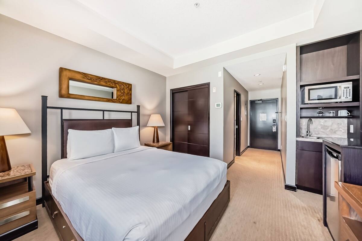 A hotel room with a large bed, bedside tables, a mirror, a wardrobe, and a kitchenette area.