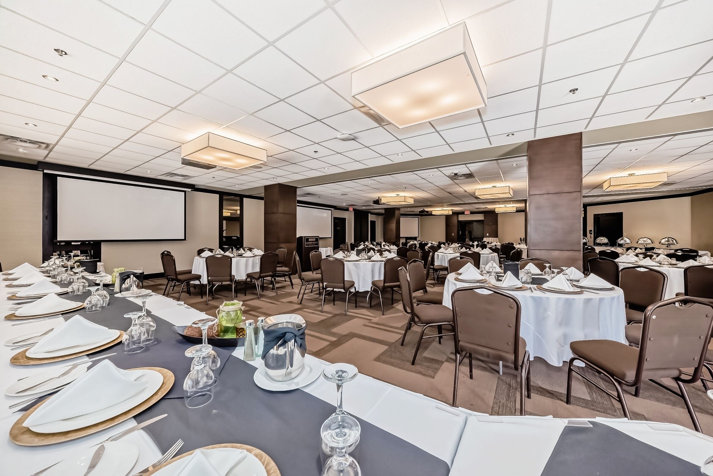 Un amplio salón de banquetes con varias mesas redondas dispuestas para comer, equipado con manteles blancos, platos, cubiertos y cristalería.