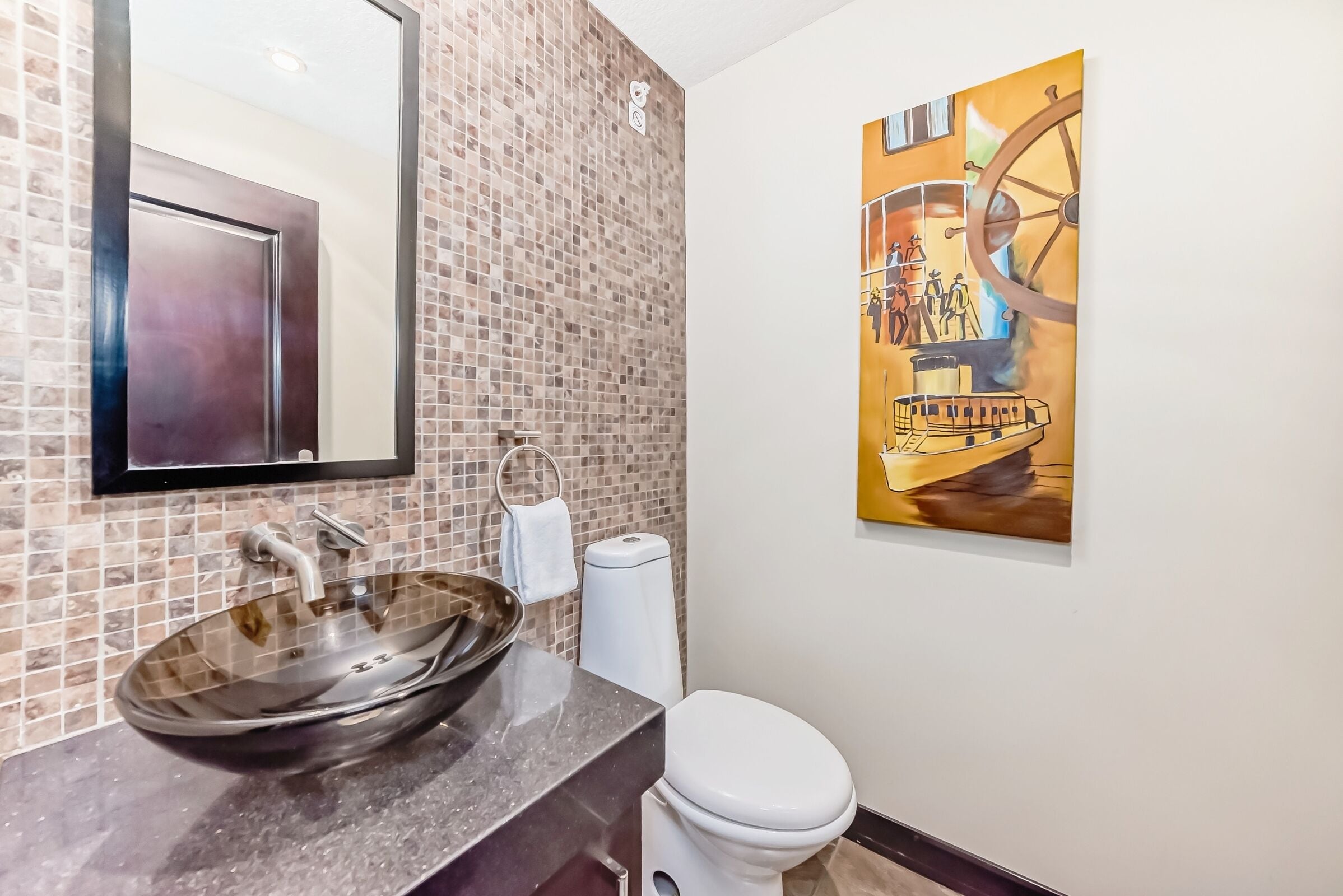 A bathroom with a mosaic tile backsplash, a large mirror, a glass vessel sink, a toilet, and a painting of a ship on a wheel.
