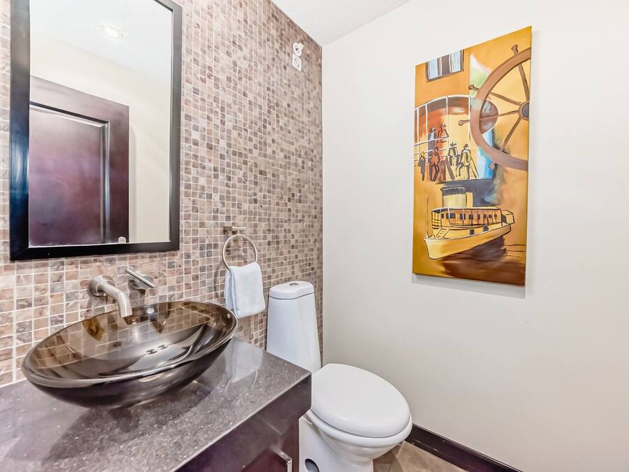 Un baño con salpicadero de mosaicos, un espejo grande, un lavabo de cristal, un inodoro y un cuadro de un barco en una rueda.