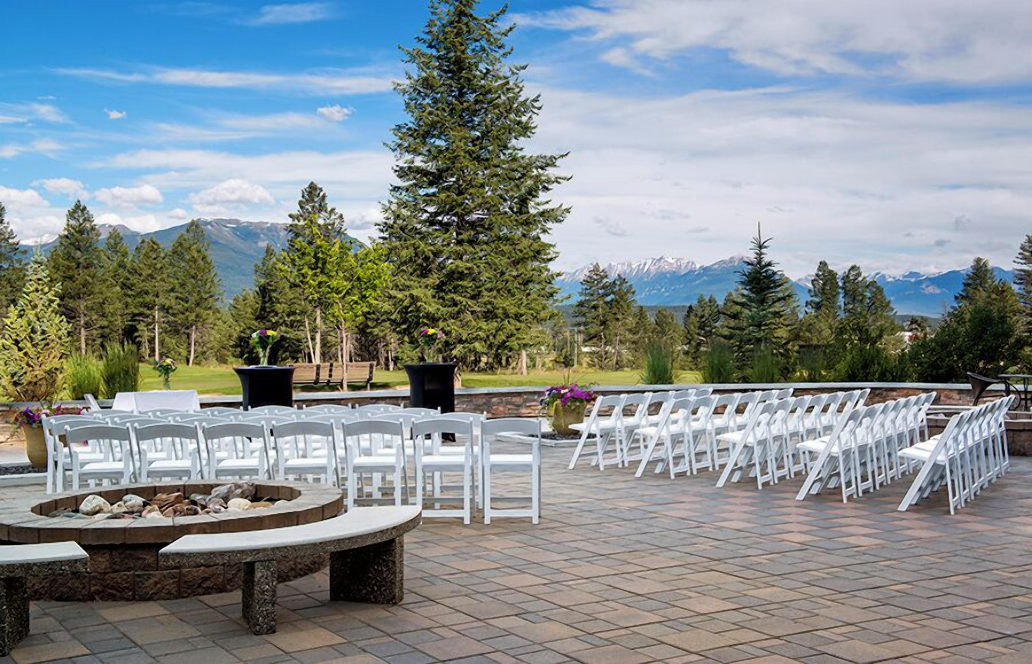 An outdoor seating area with white chairs and a stone fire pit, surrounded by trees and mountains in the background.