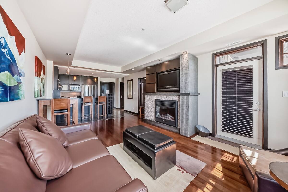 A modern living room with a brown leather sofa, a fireplace, a kitchen area with a dining table, and a large TV.