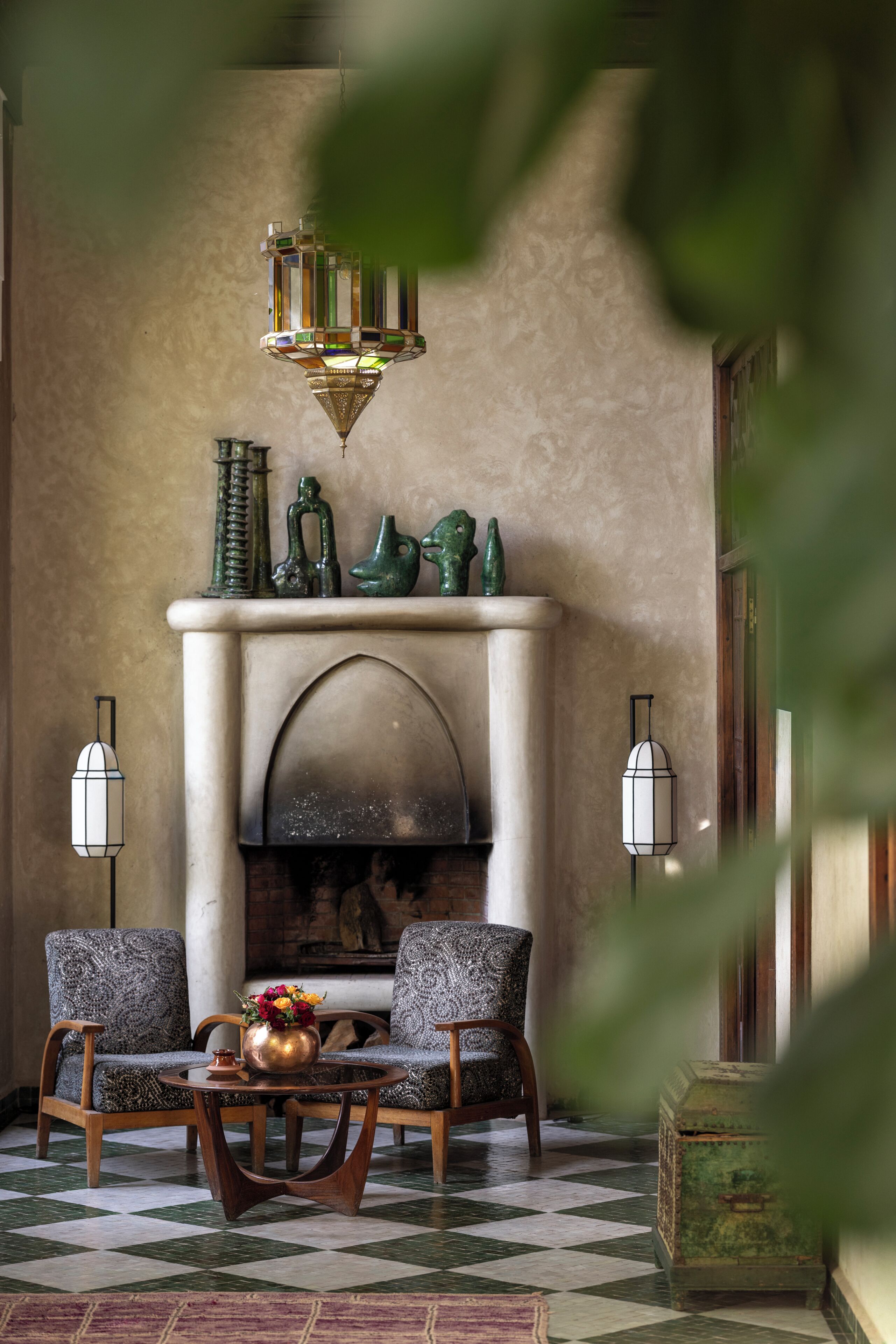 Una habitación con chimenea, dos sillones estampados, una mesa redonda de madera con un florero, y dos lámparas de pared.