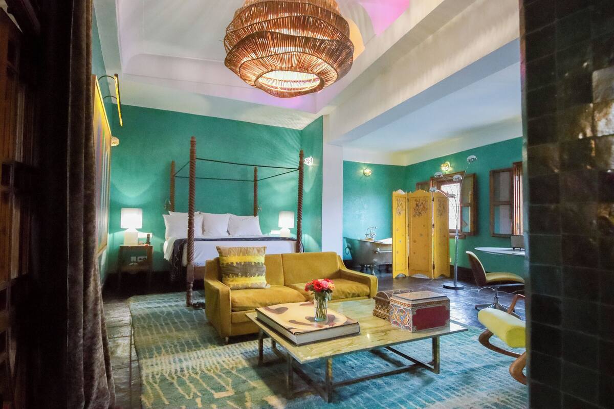 A room with a four-poster bed, a yellow armchair, a coffee table with a book and flowers, and a large wicker chandelier.