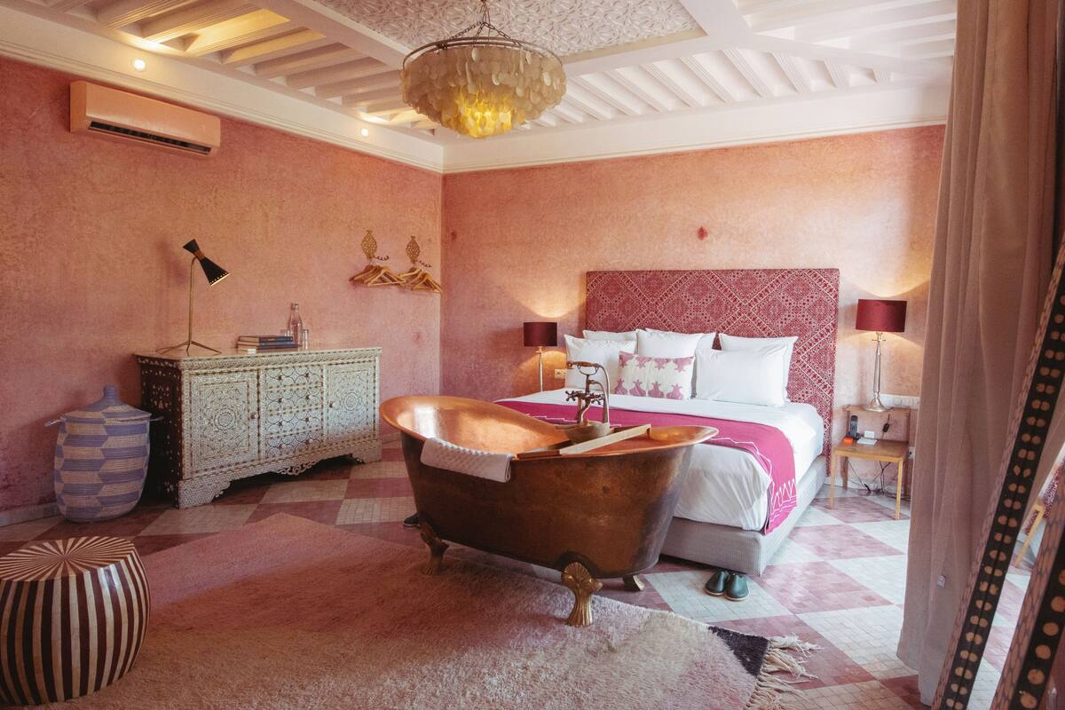 A room with a vintage bathtub, a bed with a patterned headboard, a nightstand with a lamp, and a decorative wall piece.