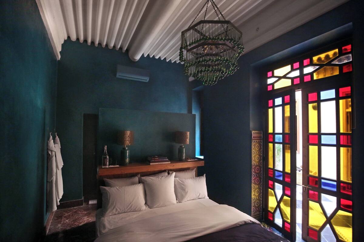 A bedroom with a bed, a wooden headboard, a hanging light fixture, and a window with colorful stained glass.