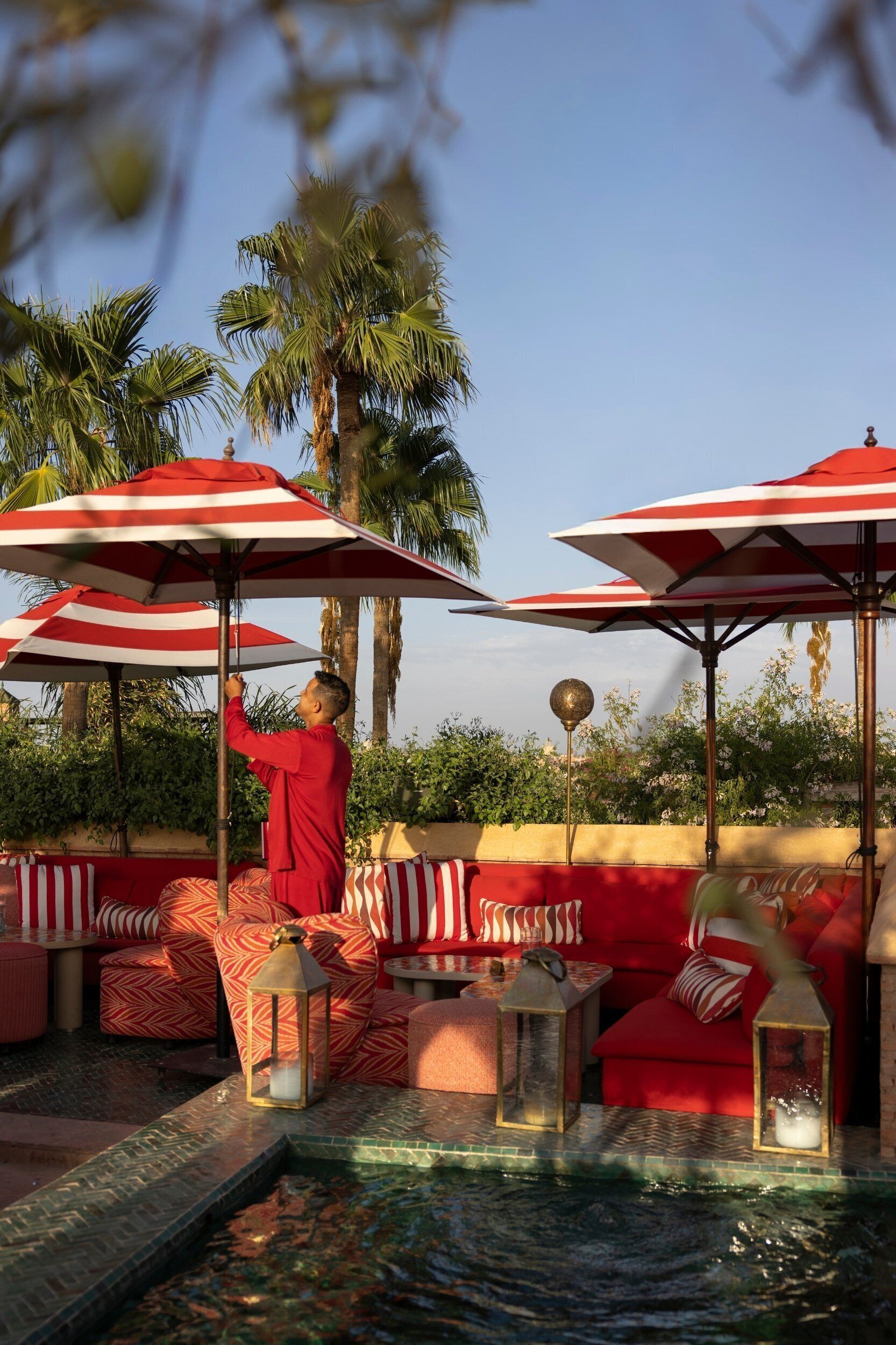 A poolside area with red and white striped umbrellas, red cushioned seating, and a water feature.