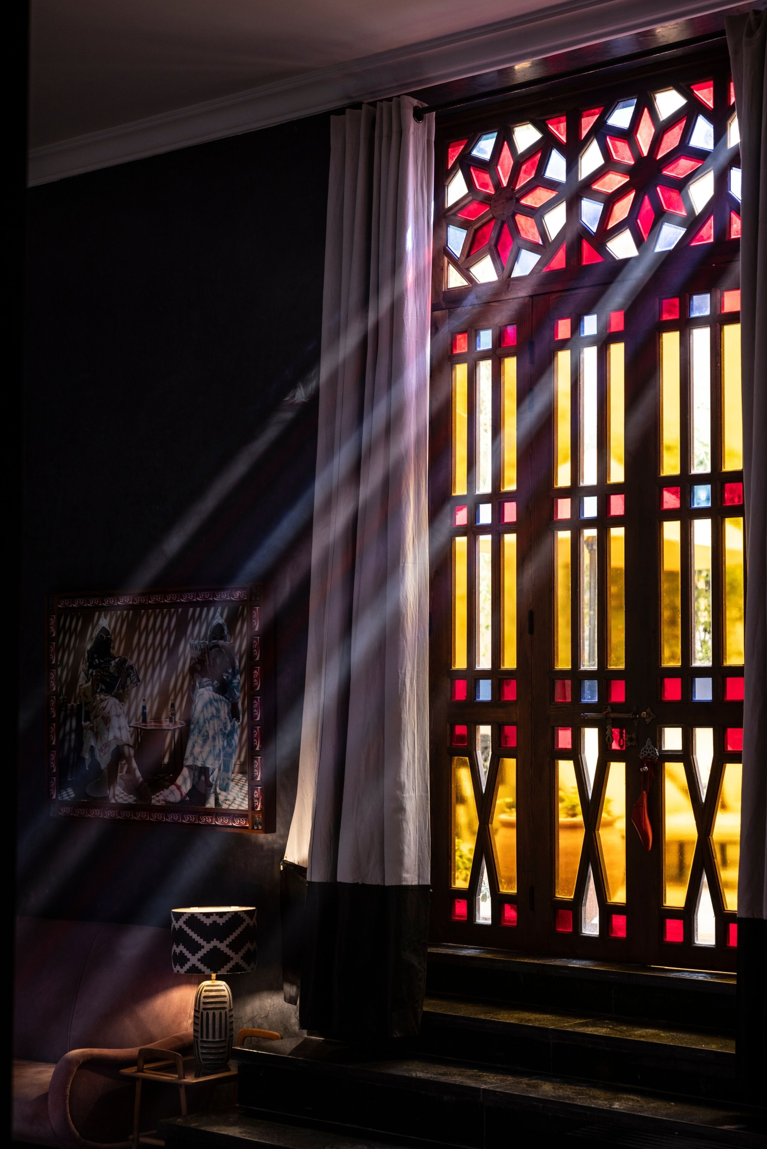 Ett rum med ett stort fönster med målat glas, ett inramat konstverk på väggen och en antik lampa.