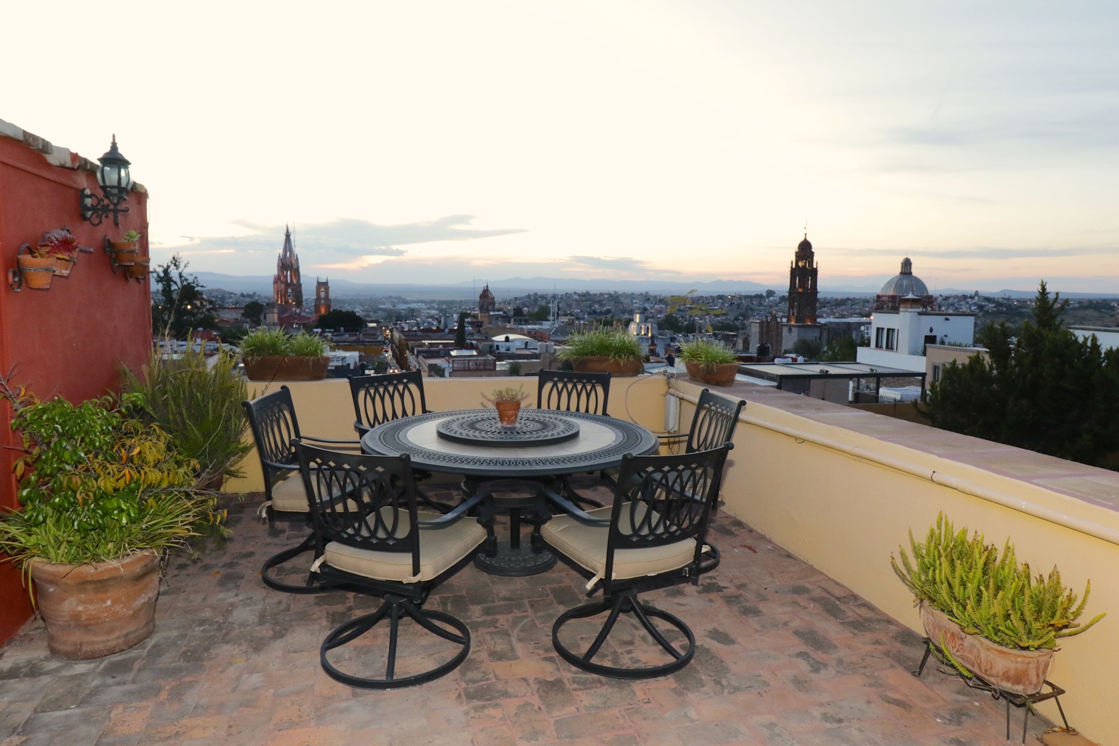 A rooftop terrace with a round table and chairs, surrounded by potted plants.
