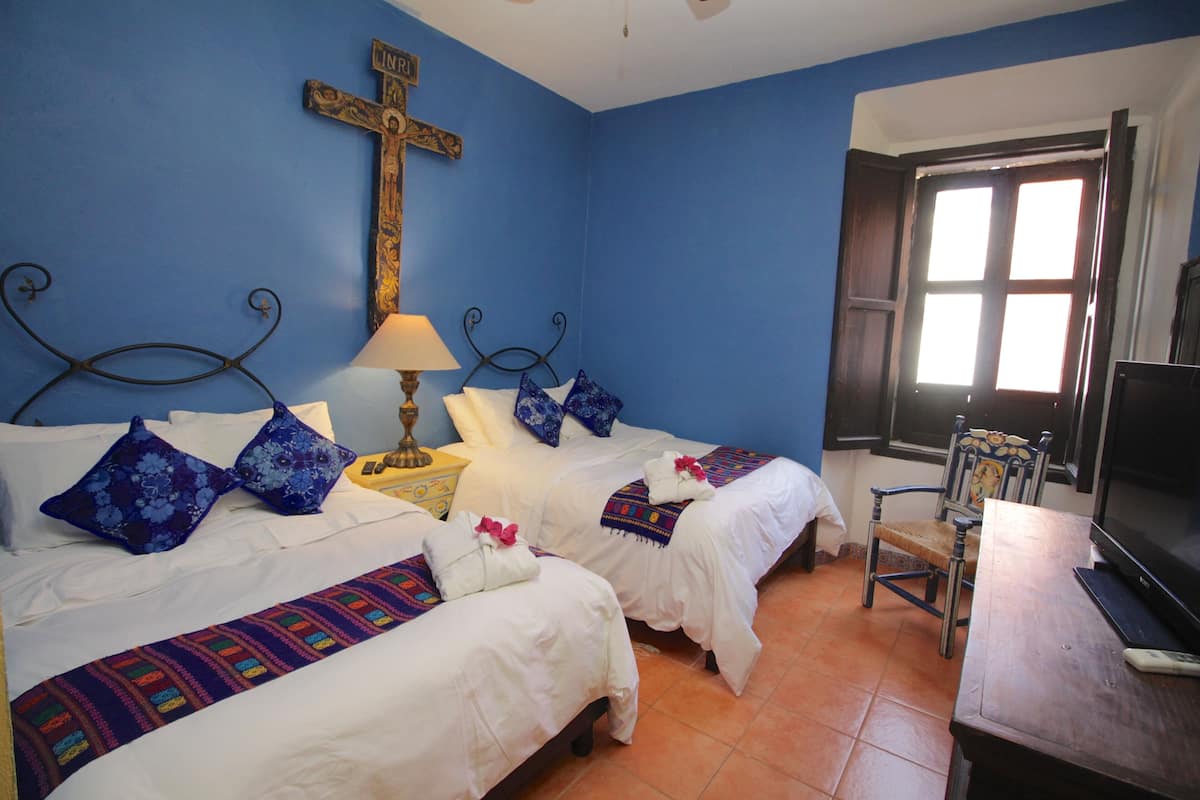 Une chambre avec deux lits, une croix au mur, une télévision et une fenêtre avec des rideaux.