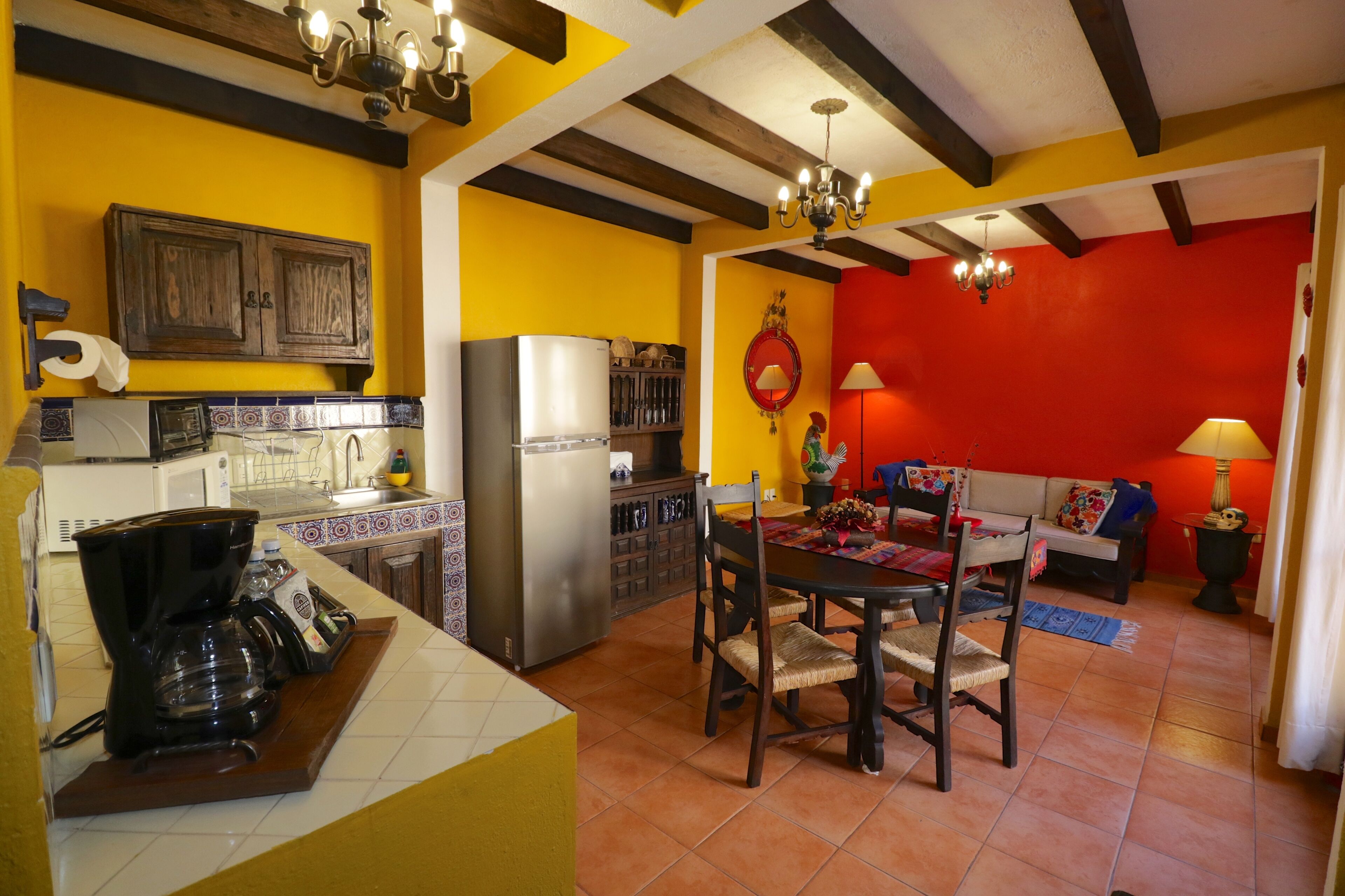 Een keuken met een koffiezetapparaat, een koelkast en houten kastjes. Er staat een eettafel met stoelen, en achtergrond is een rode muur.