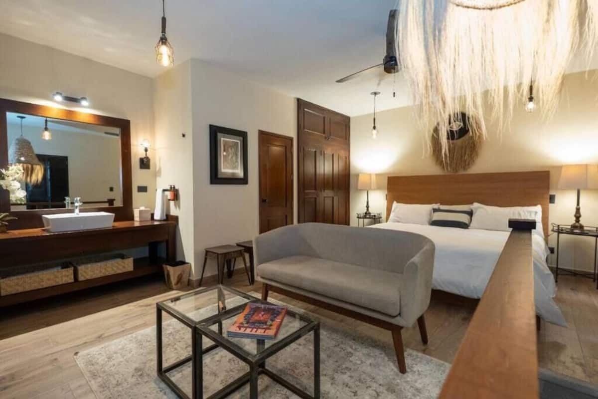 Une chambre d’hôtel moderne avec un grand lit, un canapé, une table basse en verre, une table d’appoint en bois, une coiffeuse avec un miroir et un ventilateur de plafond.