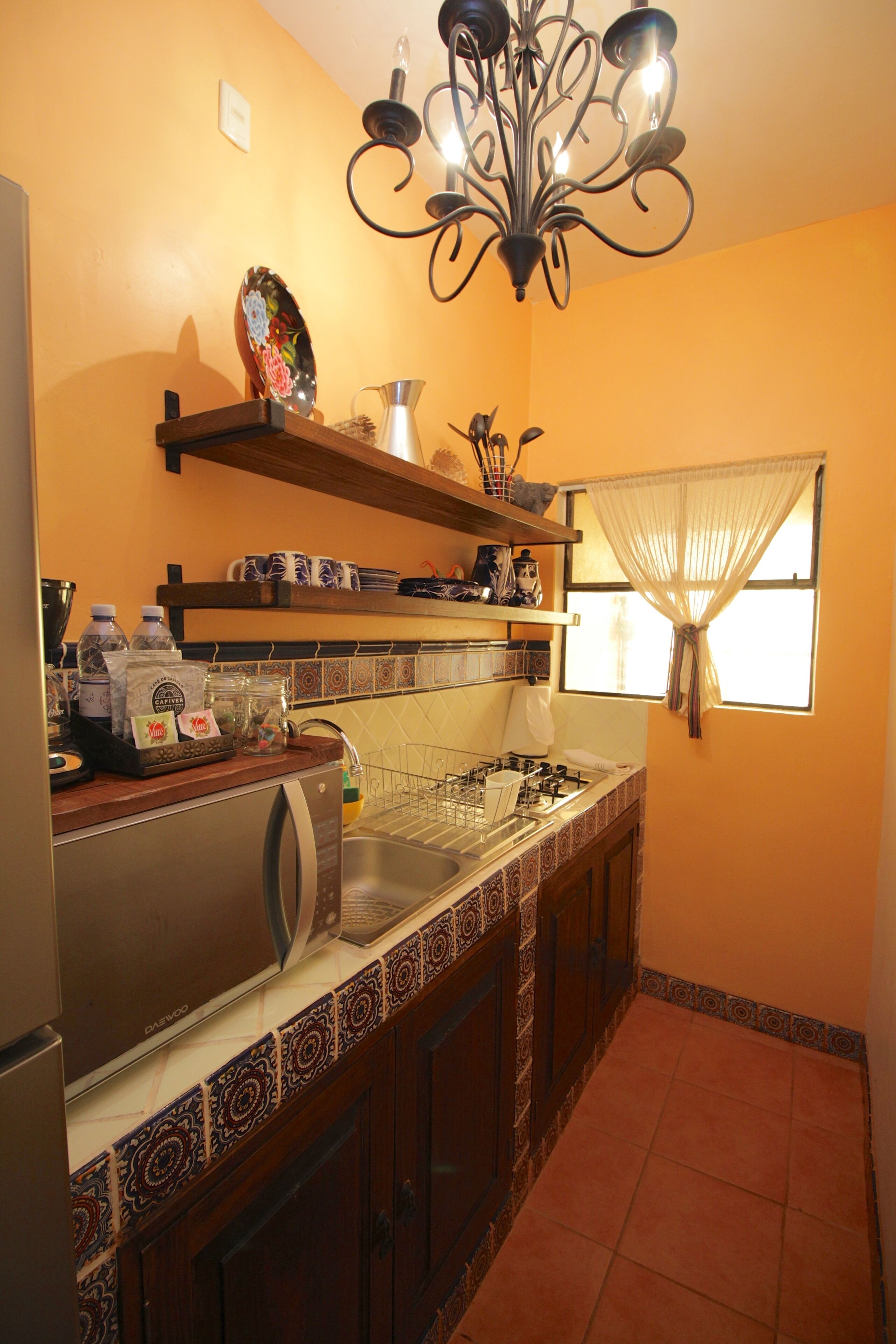 A compact kitchen with a refrigerator, stove, and sink. There are shelves above the counter holding various kitchen items, and a window with a curtain.