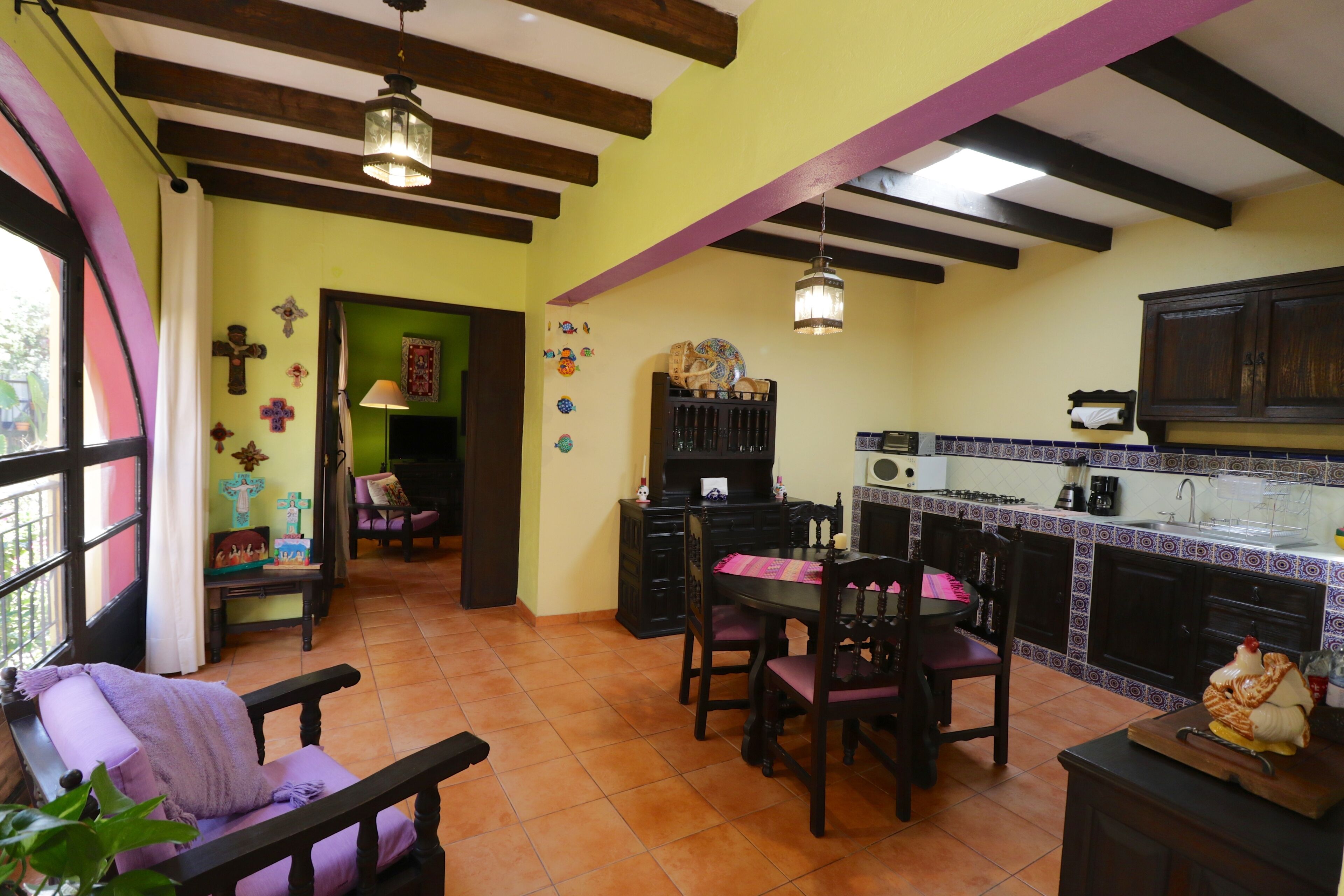 A cozy kitchen with a dining area, featuring wooden beams, a tiled floor, and a window with curtains.