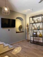 Una sala de estar con televisor, piso de madera, estante metálico con adornos y una lámpara colgante.