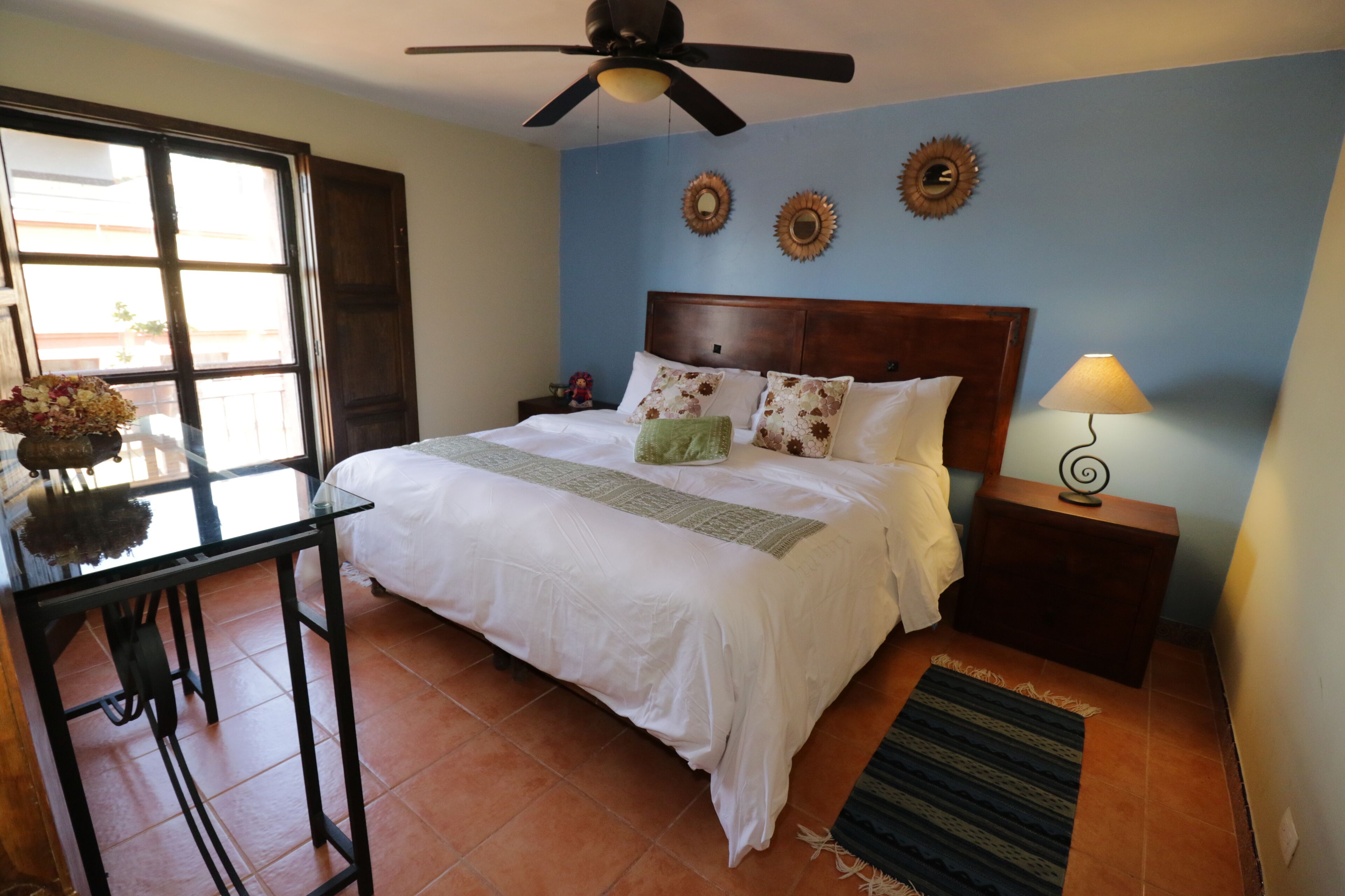 A bedroom with a bed, a nightstand, a lamp, a ceiling fan, a window, and a table with a vase of flowers.