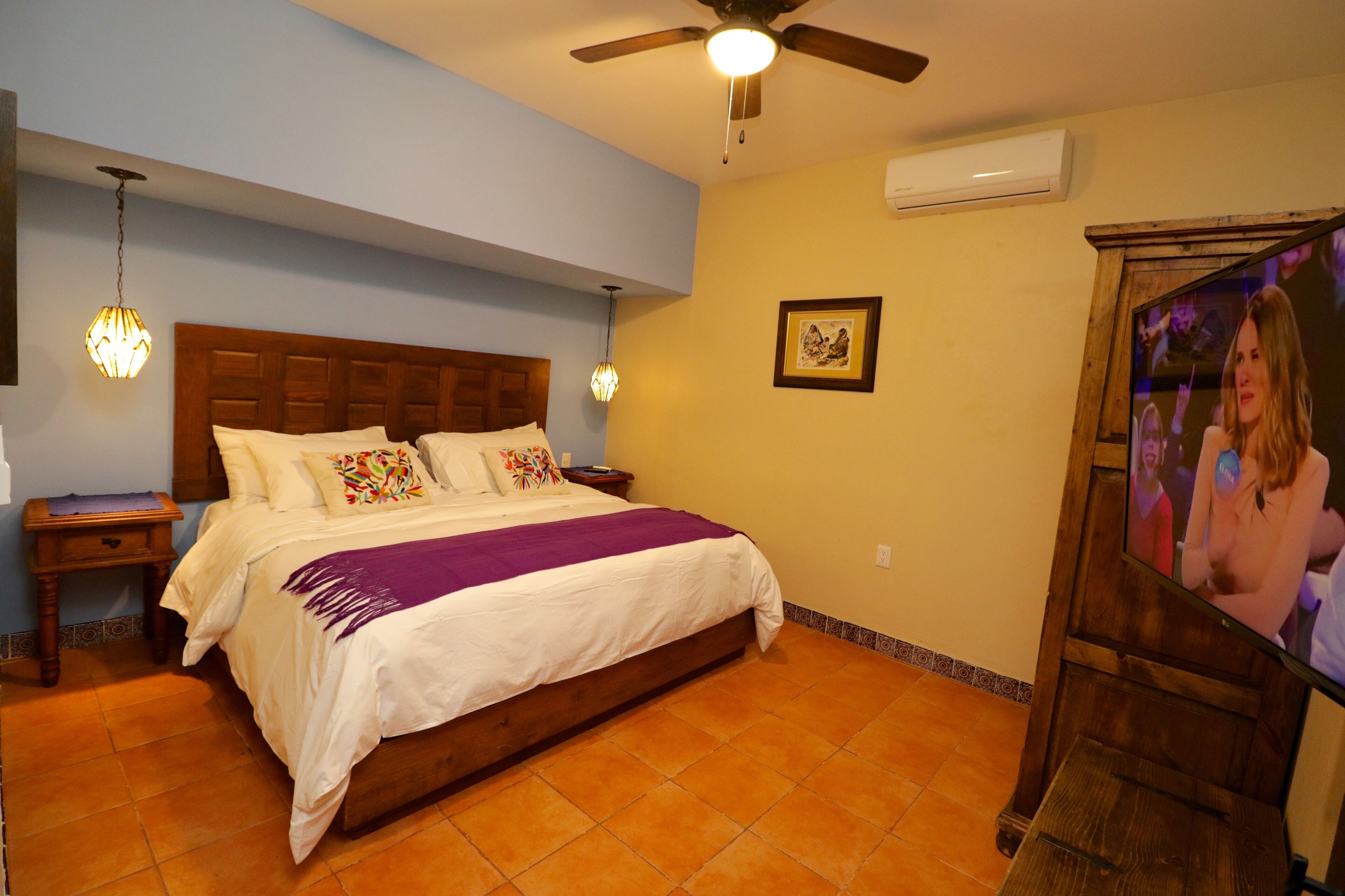 Un dormitorio con cama, cabecera de madera, mesitas de noche, ventilador de techo y televisión.