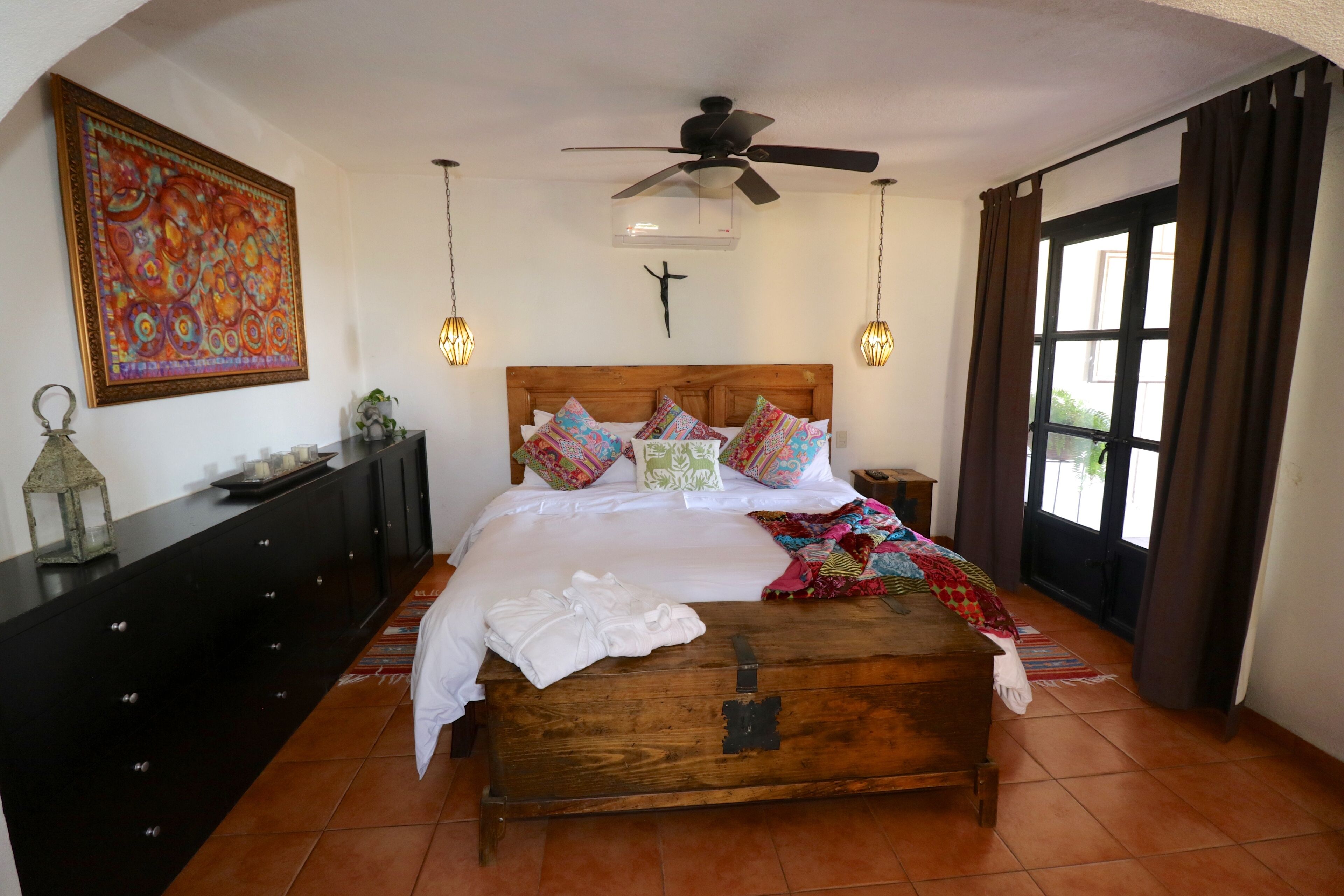 Un dormitorio con cama, un baúl de madera, ventilador de techo, aire acondicionado montado en la pared y una ventana con cortinas.
