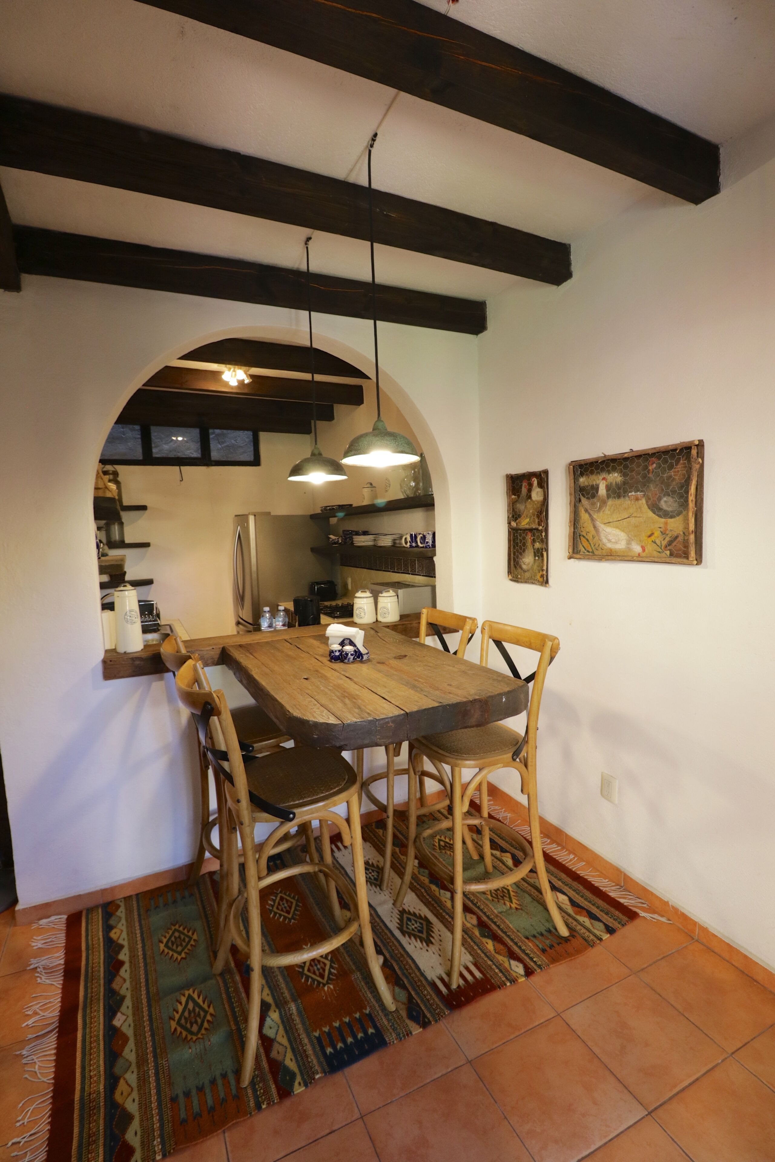 A dining area with a wooden table and chairs, a patterned rug, and a kitchen in the background.
