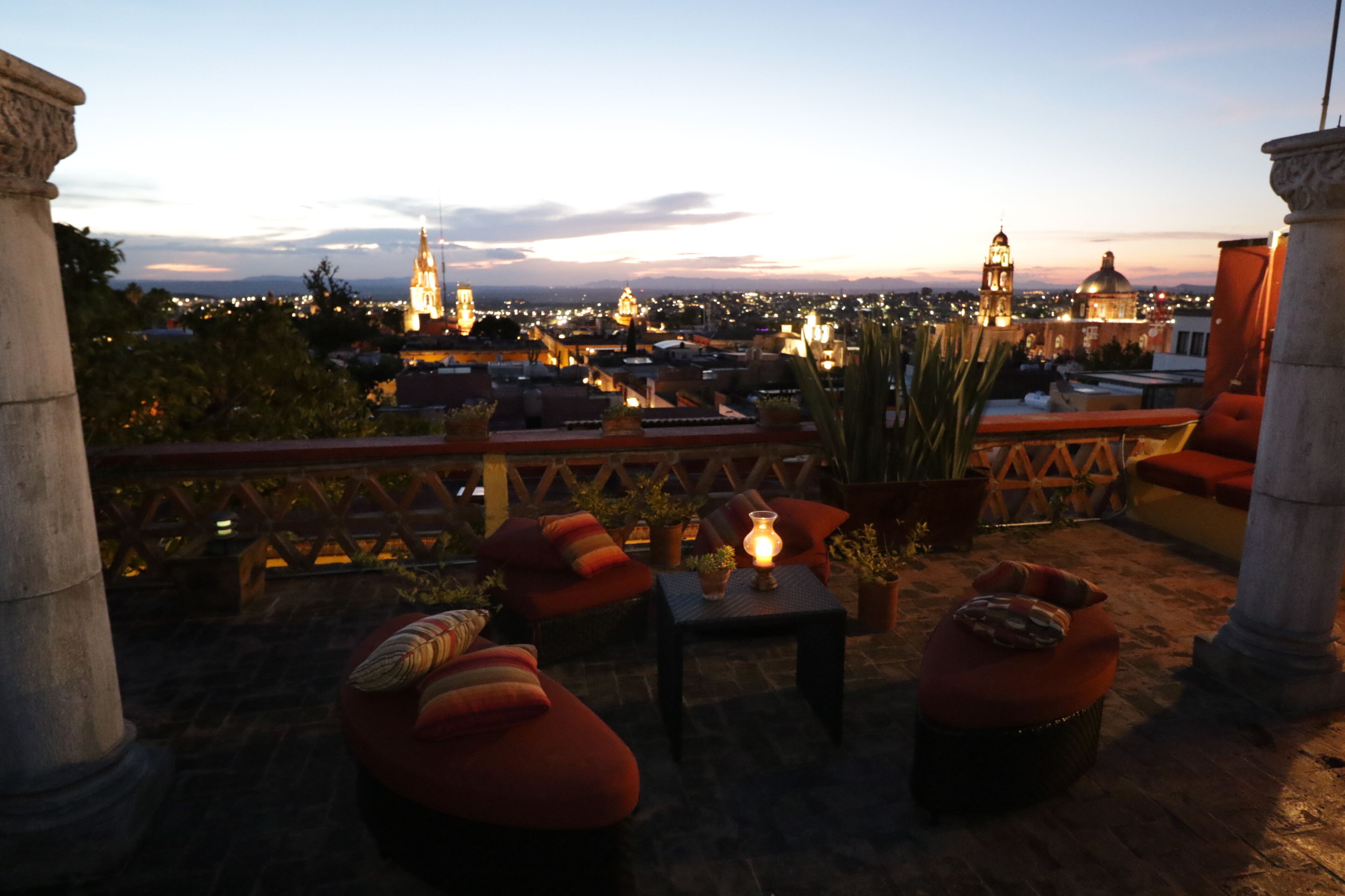 A rooftop terrace with outdoor seating, a table with a lamp, and a view of a cityscape at dusk.