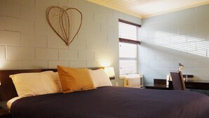 Une chambre avec un grand lit, un décor en forme de cœur sur le mur, un bureau avec une chaise et une fenêtre avec des stores.