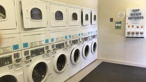 Une laverie équipée de plusieurs machines à laver et de plusieurs séchoirs, de panneaux de commande et d’un panneau indiquant où trouver le savon, l’eau de Javel et l’adoucissant.