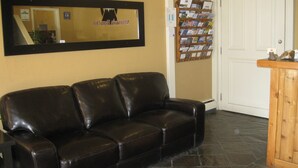 Un canapé en cuir noir se trouve dans un hall d’entrée, agrémenté d’un miroir encadré, d’un panneau d’affichage où sont accrochées des brochures et d’un comptoir de réception en bois.