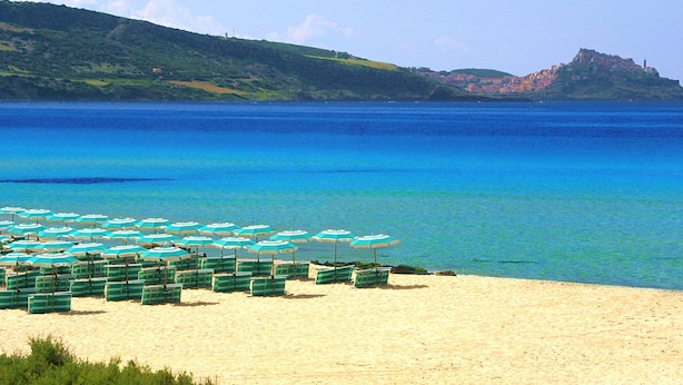 Ein Strand mit Reihen aus grün-weißen Liegestühlen und Sonnenschirmen, ein sandiger Uferstreifen und eine in der Ferne gelegene Hügel mit Gebäuden.