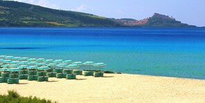 A beach with rows of green and white beach chairs and umbrellas, a sandy shore, and a distant hill with buildings.