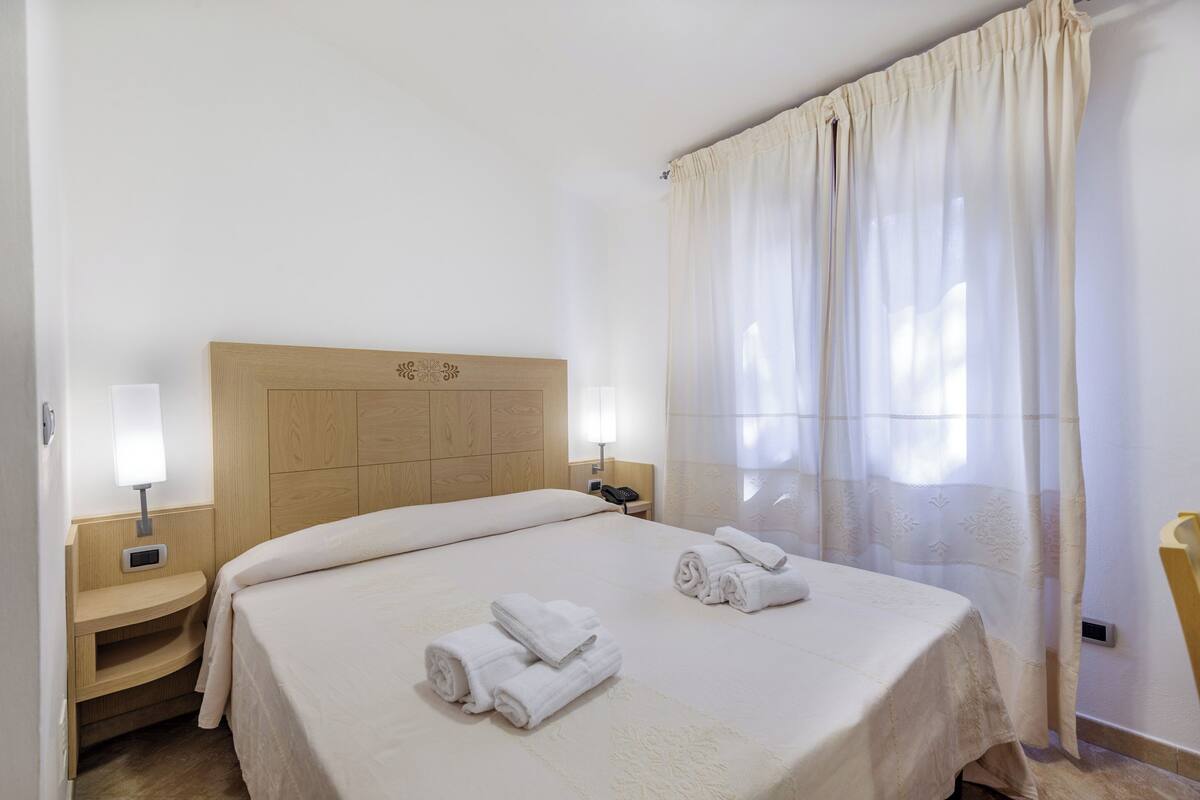Una camera d'albergo con una testiera in legno, due comodini con lampade, un letto con copripiumino bianco e una finestra con tende leggere.
