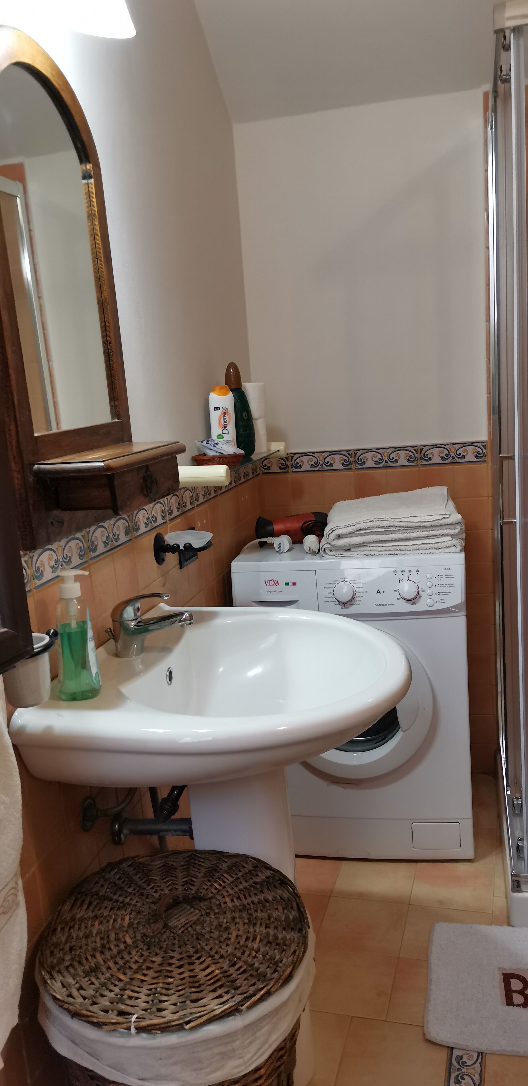 Un baño con lavamanos blanco, espejo, lavadora y cesta de mimbre.