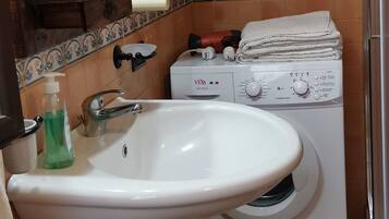 Un baño con lavamanos blanco, espejo, lavadora y cesta de mimbre.