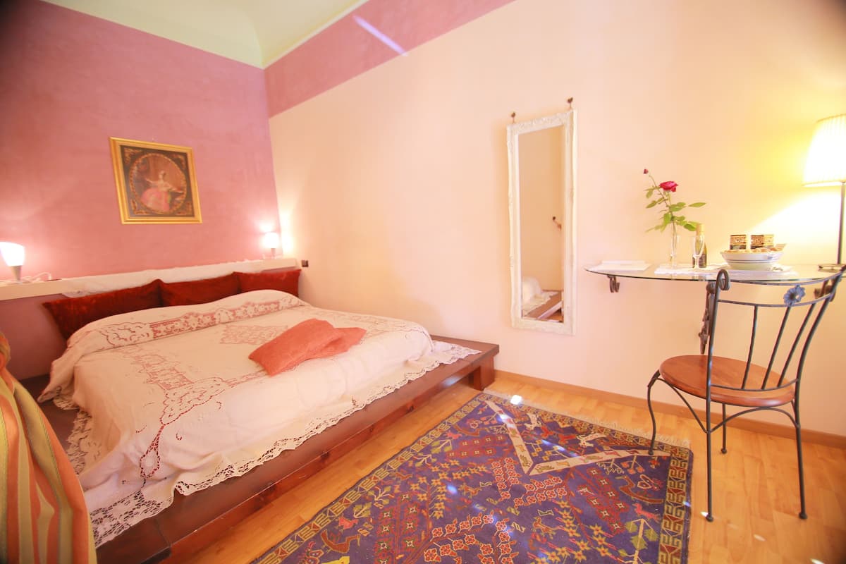 Ein Schlafzimmer mit einem Bett, einem Sessel, einem kleinen Tisch mit einer Vase und einem Spiegel an der Wand.