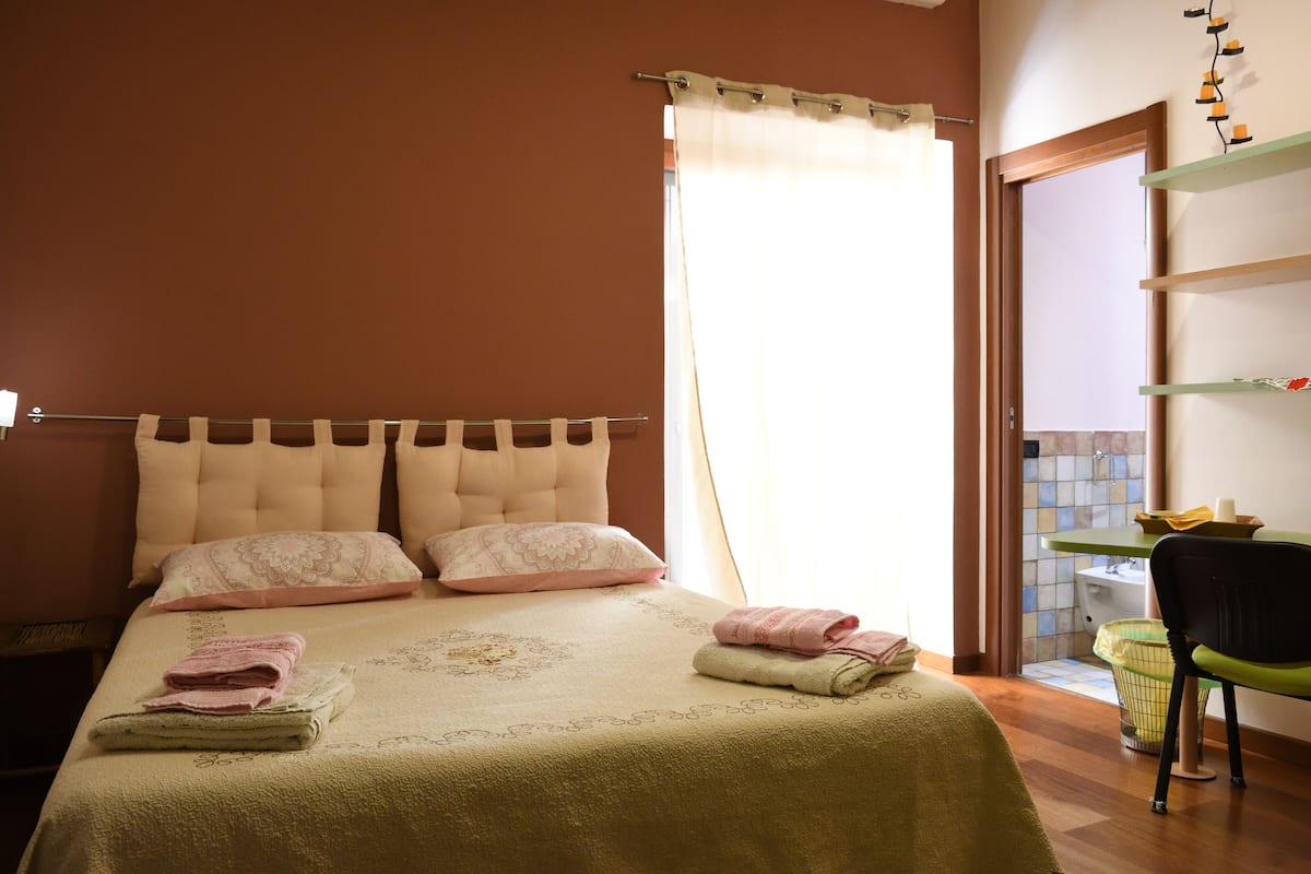 Ein ordentlich gemachtes Bett mit Kissen und gefalteten Handtüchern, ein Nachttisch mit Lampe und ein Stuhl in der Nähe eines Fensters mit Vorhängen.